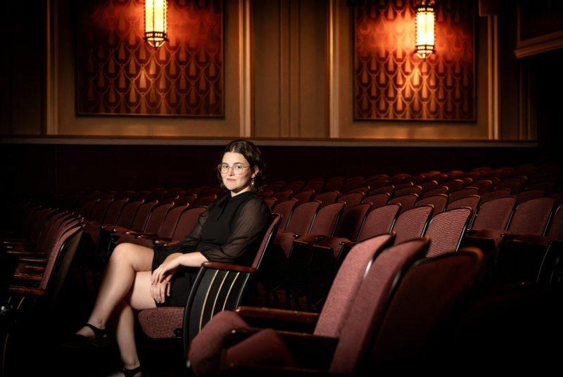 A woman sits on a seat within a row of seats in a theatre.